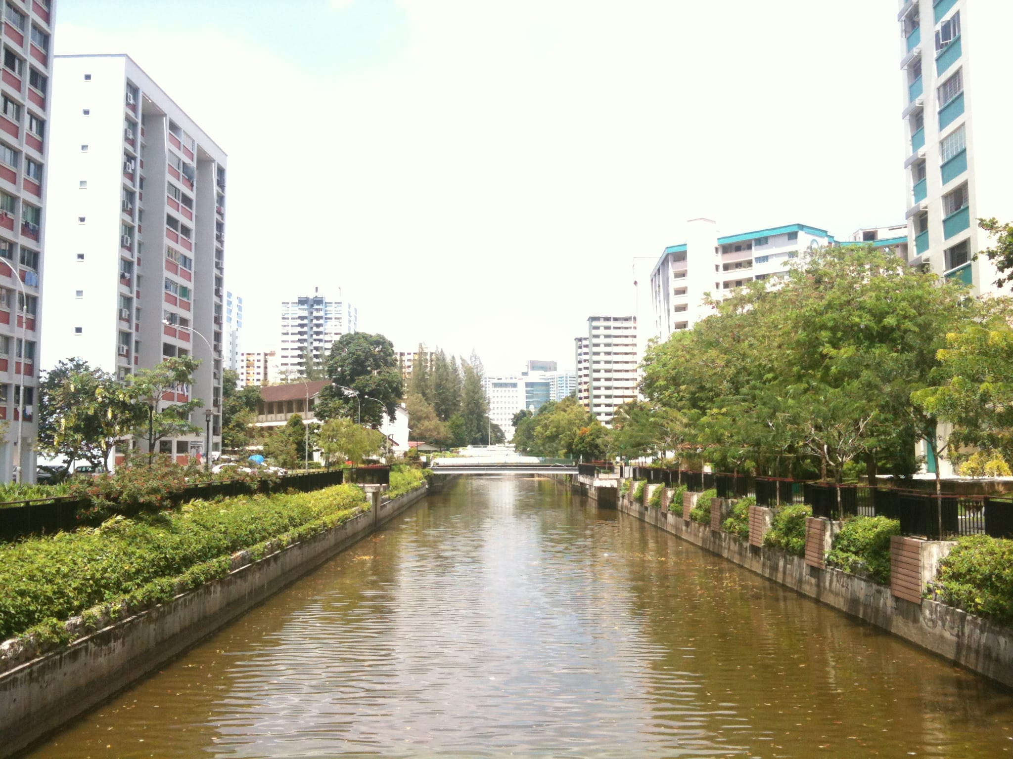 A canal is flanked by tall apartment buildings and lush greenery on both sides, with a bridge in the distance.